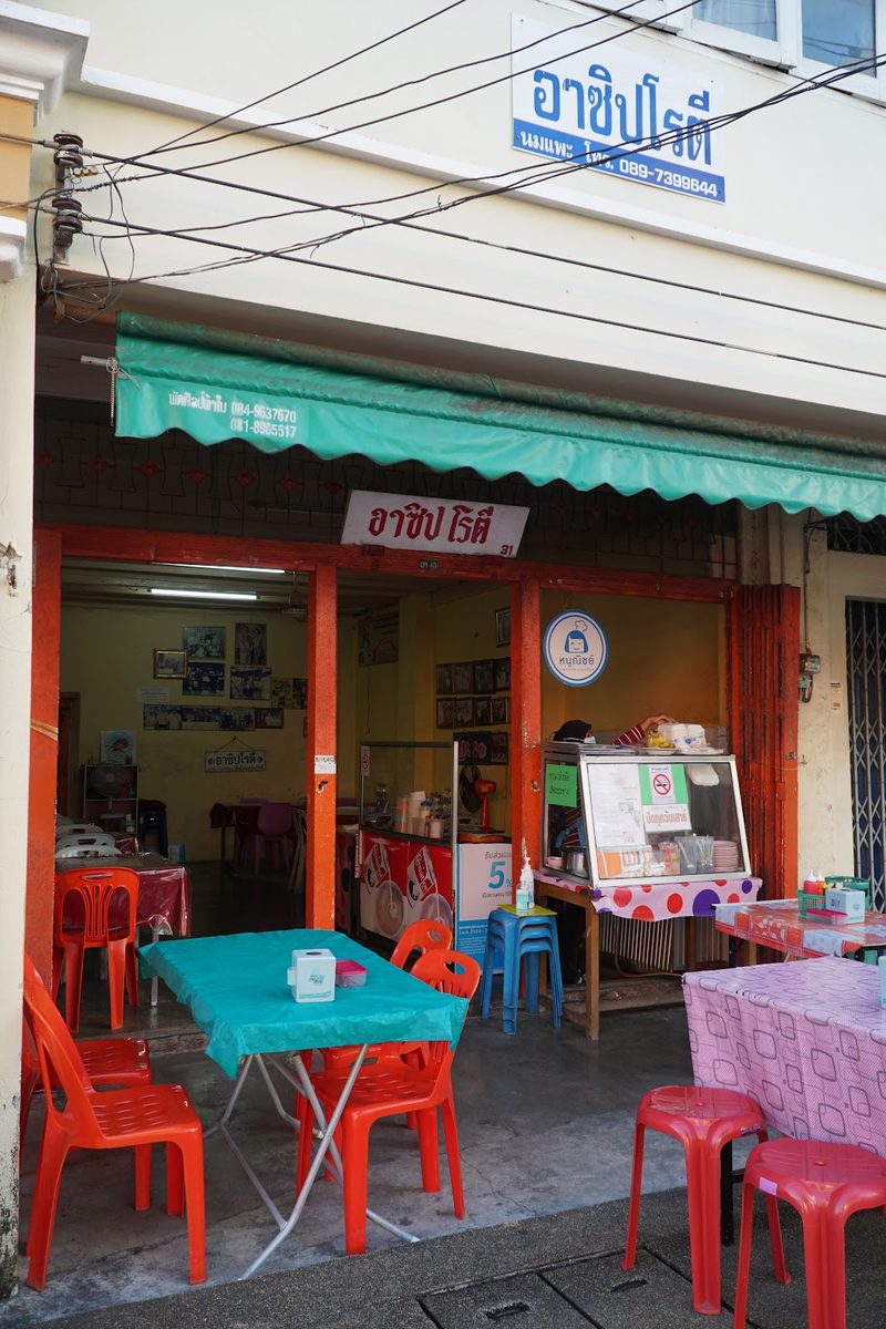 ตึกแถวหน้าร้าน ป้ายชื่อร้านอยู่เหนือกันสาดสีเขียว มีโต๊ะเก้าอี้วางริมทางเดิน